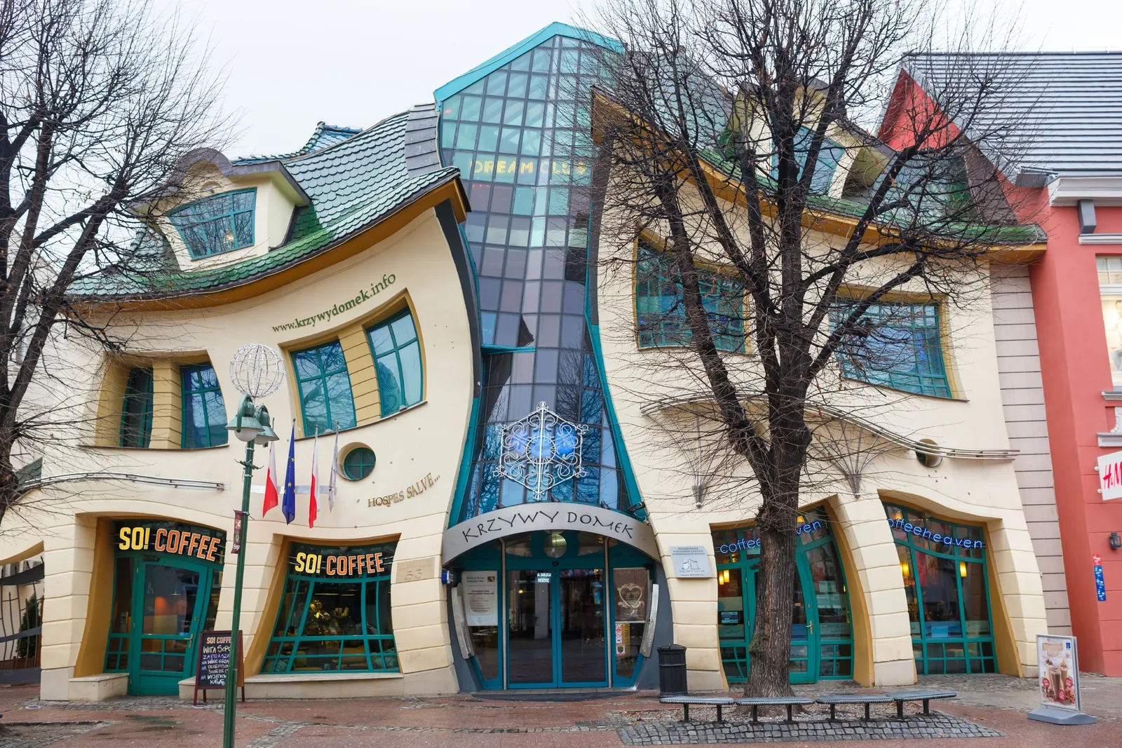 A whimsical, distorted building with curved walls and windows, known as the Crooked House (Krzywy Domek), featuring cafes and shops at street level, with leafless trees in front.