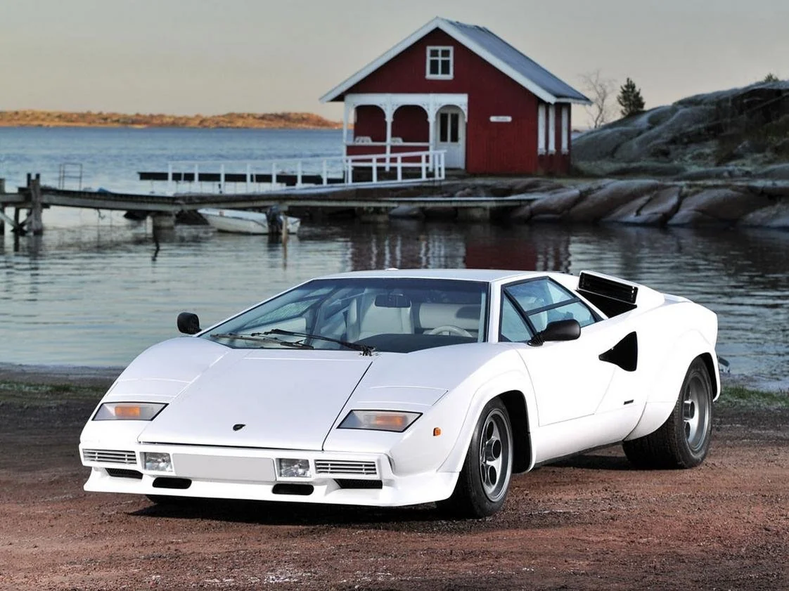 A white Lamborghini Countach sports car is parked on a dirt road near a body of water, with a small red and white house and a dock in the background.