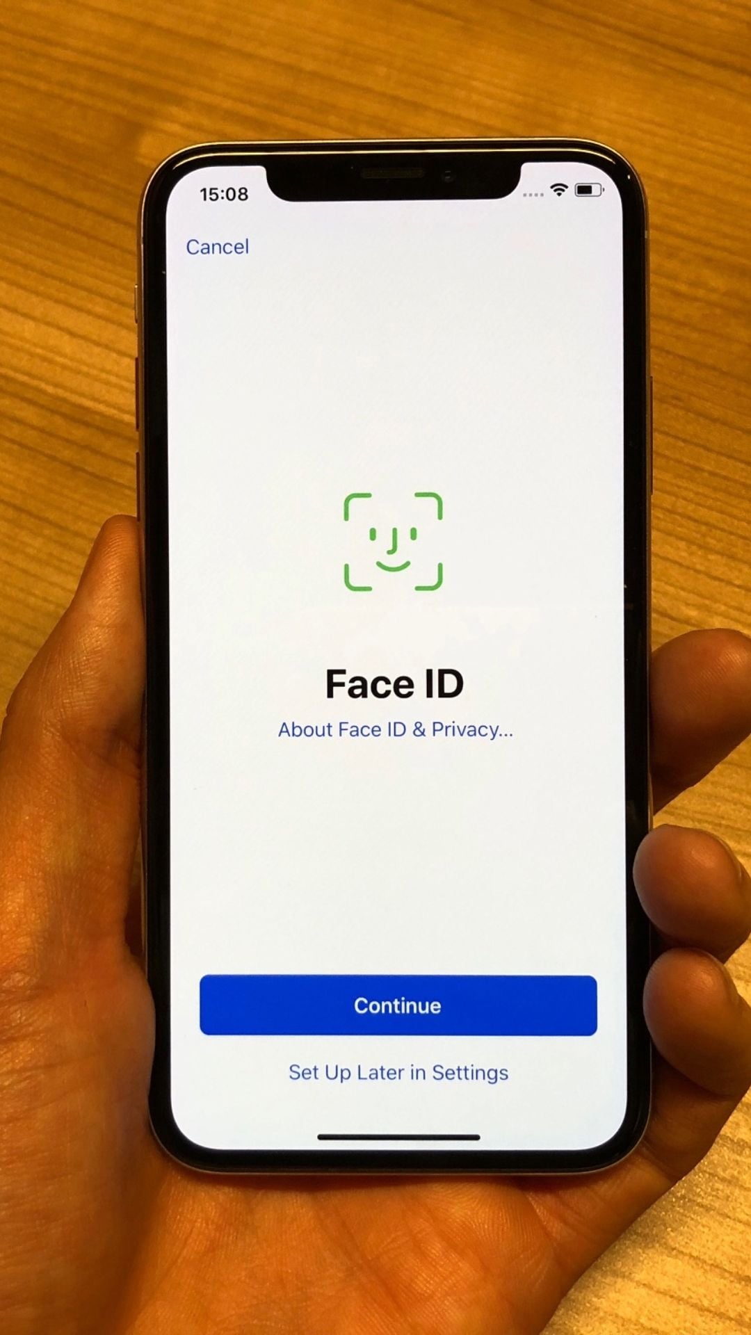 A hand holds a smartphone displaying the Face ID setup screen, with options to continue or set up later. The phone is against a wooden surface background.