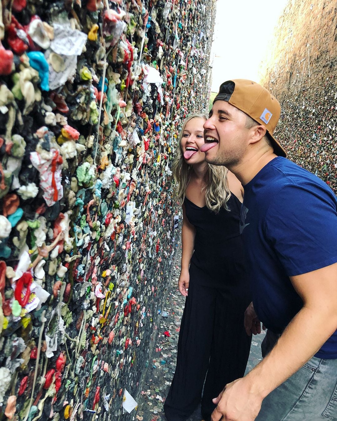 A woman and a man wearing casual clothes lean toward a wall covered in colorful pieces of chewing gum, playfully sticking out their tongues as if about to lick the gum wall.