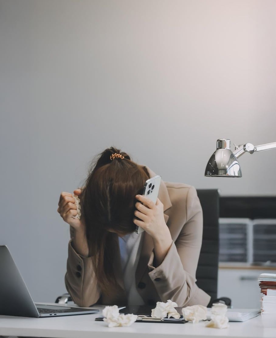 A woman sits at a desk with her head in one hand and a phone in the other, looking stressed or frustrated. Crumpled papers are scattered on the desk, next to a laptop and a lamp.