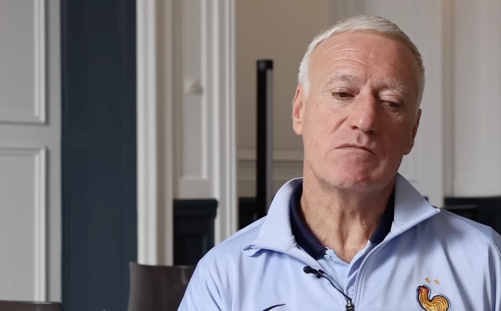 A man with short gray hair wearing a light blue sports jacket with a rooster emblem sits indoors, looking thoughtful. The background features white paneled walls and some dark furniture.