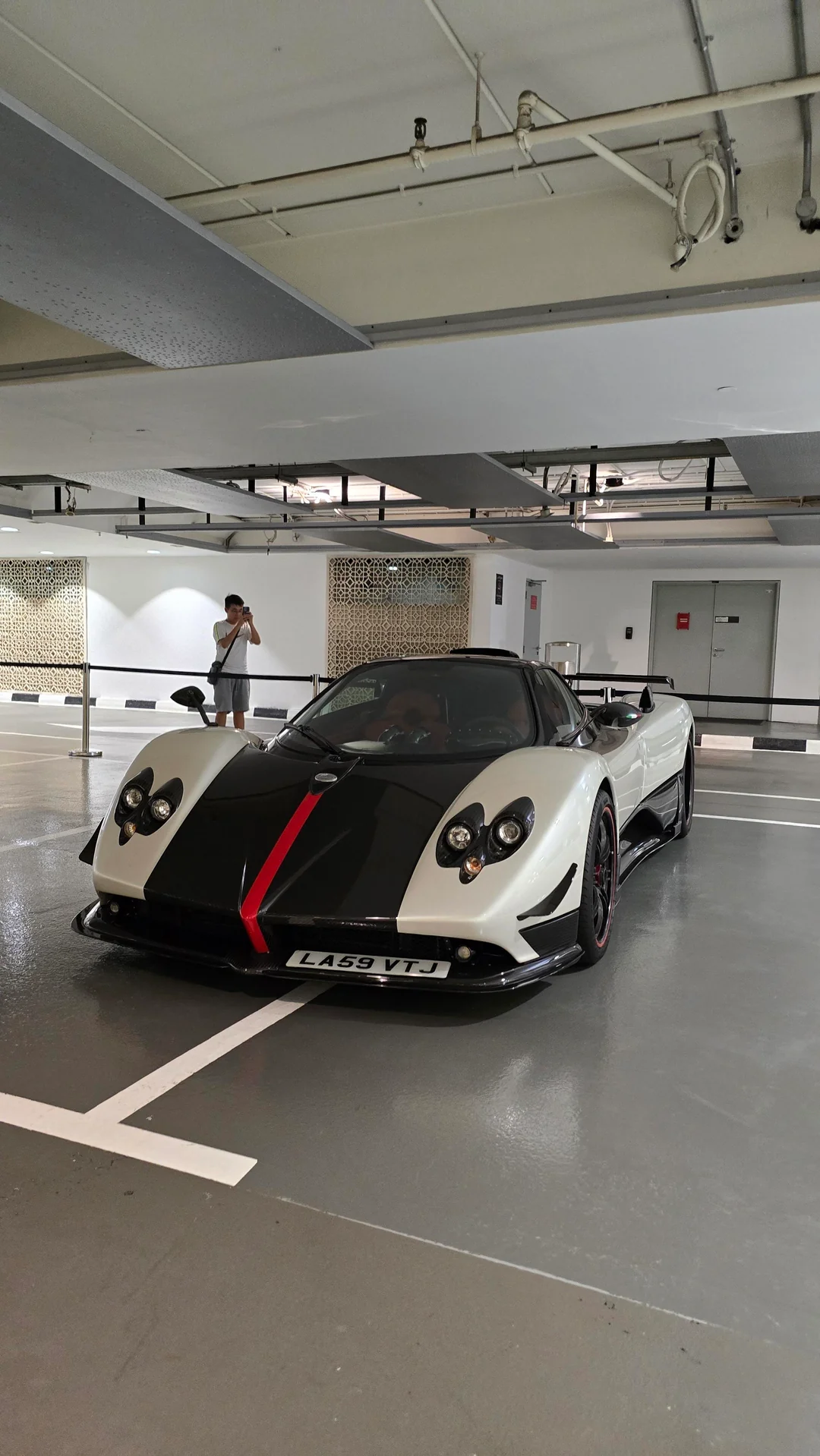 A white and black sports car with a red stripe is parked in an indoor garage. A person in the background is taking a photo of the car with a phone. The setting is well-lit with gray flooring and exposed ceiling pipes.