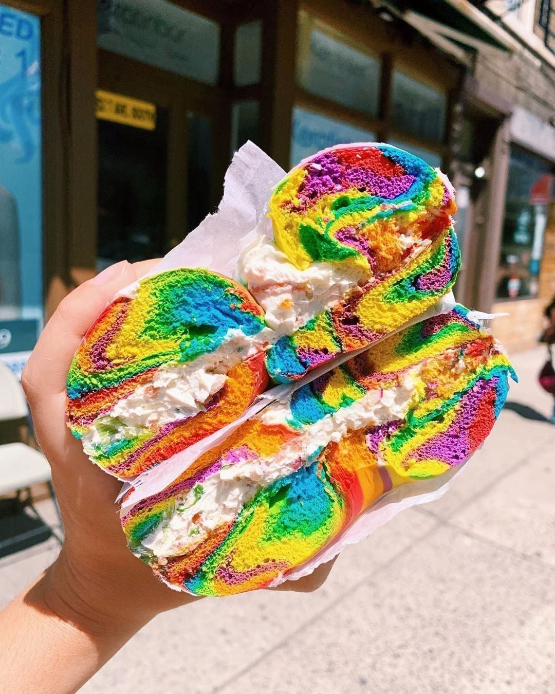 A hand holds a rainbow-colored bagel sliced in half, filled with cream cheese, against a sunny outdoor street background.