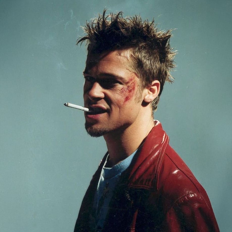 A man with messy spiked hair, facial bruises, and a cigarette in his mouth wears a red leather jacket and looks to the side against a plain background.