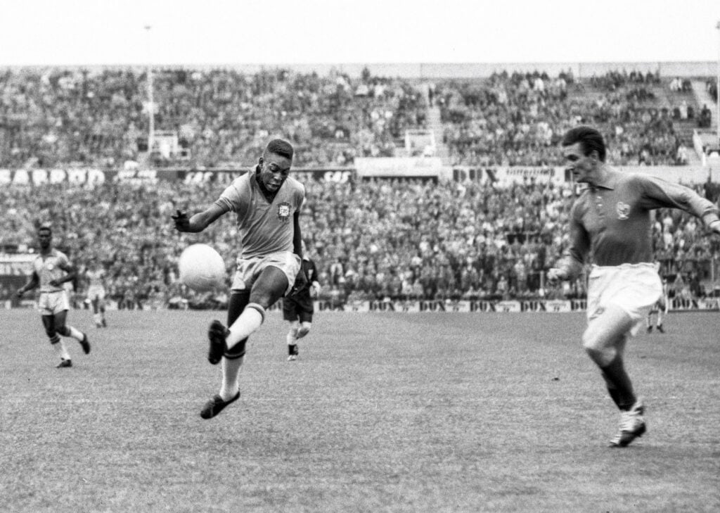 A black-and-white photo of a soccer match shows a Brazilian player kicking the ball mid-air while an opposing player approaches. The stadium is full of spectators in the background.
