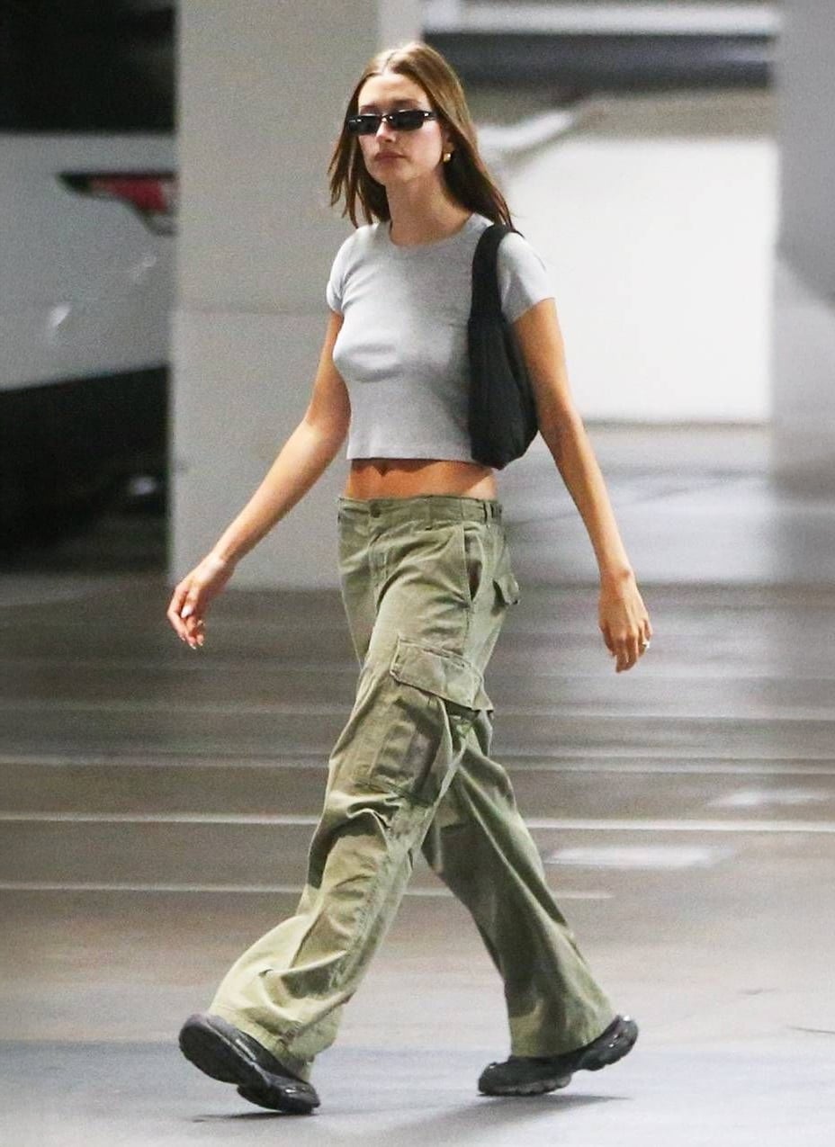 A woman in a grey crop top, loose olive cargo pants, black shoes, sunglasses, and a black shoulder bag walks in a parking garage.