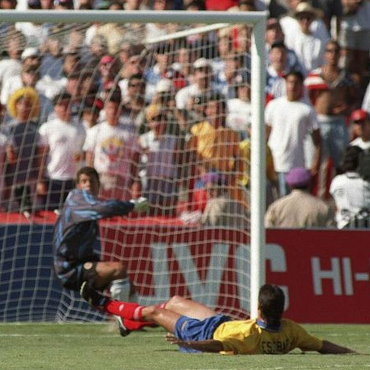 A soccer goalkeeper dives to his left as a player in a yellow jersey slides on the grass near the goalpost, with a crowd of spectators watching in the background.