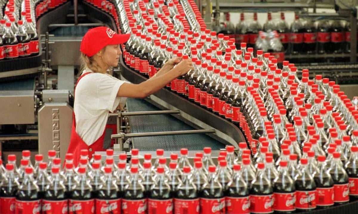 A worker in a red Coca-Cola cap and uniform inspects rows of Coca-Cola bottles moving along a conveyor belt in a bottling factory. Hundreds of bottles are lined up in front of her.