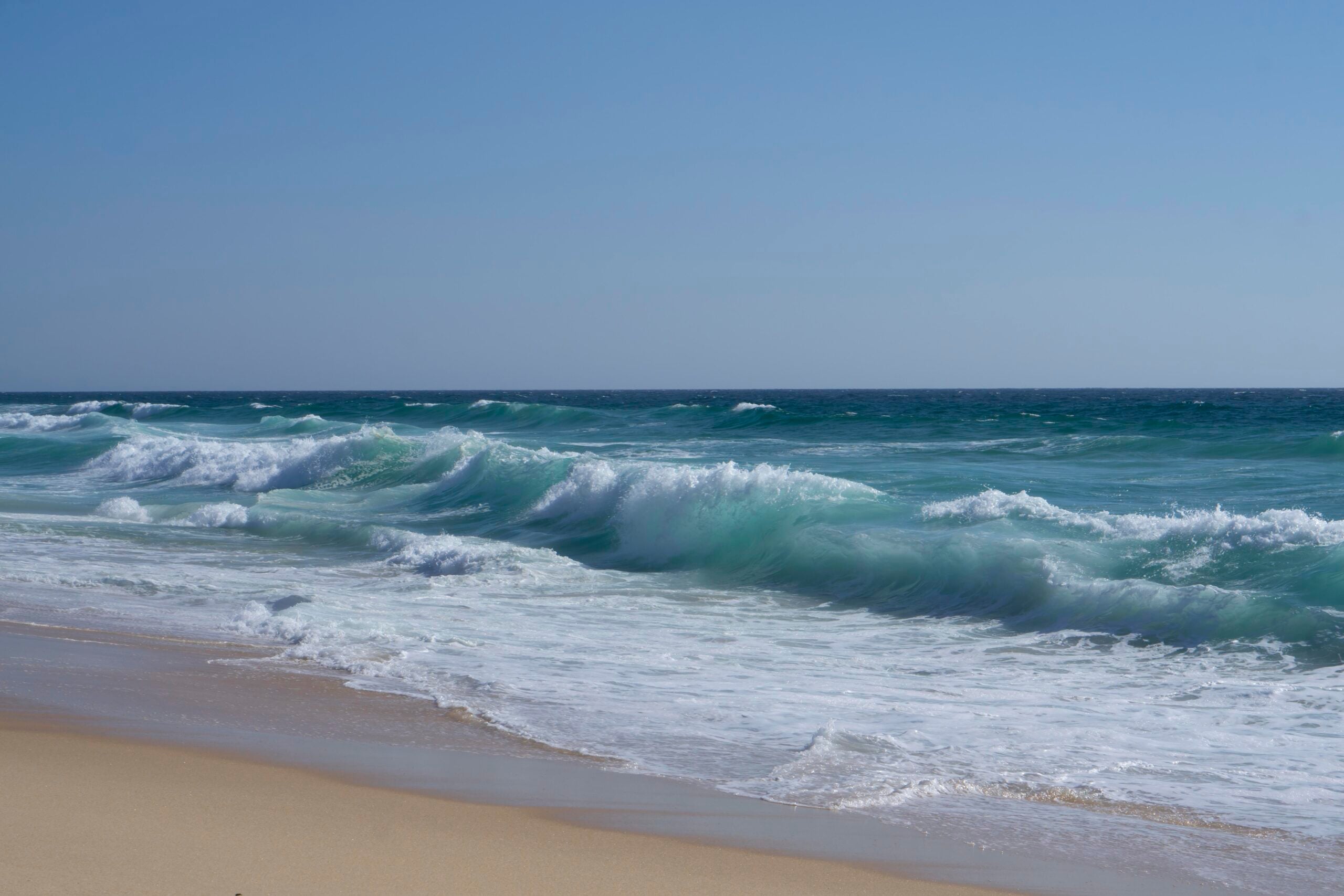 Waves crash onto a sandy beach under a clear blue sky, with turquoise and deep blue water stretching to the horizon.