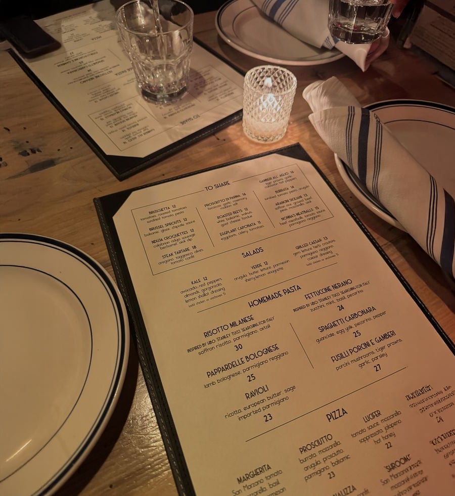 A restaurant table set with plates, glasses, a candle, and a cloth napkin. A menu featuring salads, pasta, and pizza options is in focus, with another menu partially visible in the background.