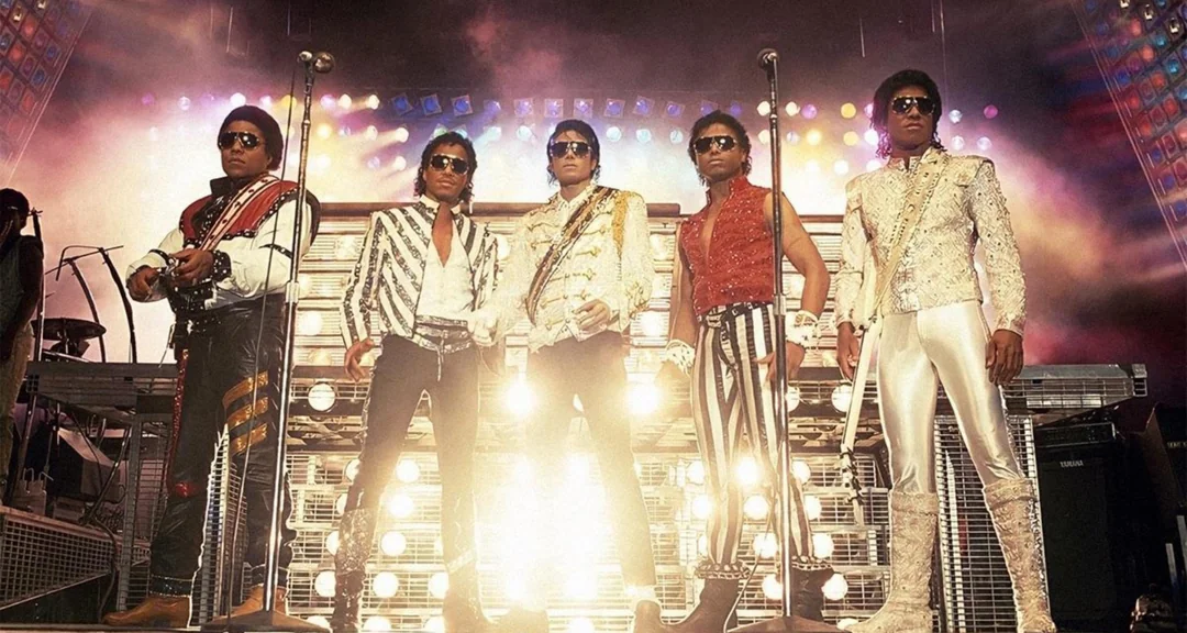 Five men in elaborate, colorful costumes and sunglasses stand on a brightly lit concert stage, holding microphones, with dramatic lighting and musical equipment in the background.