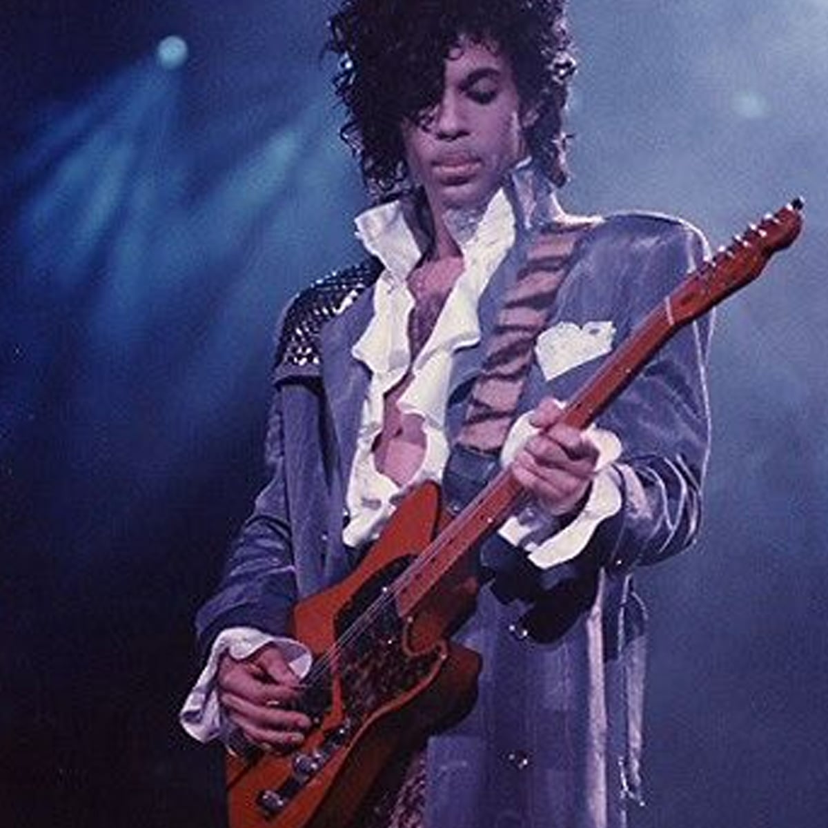 A musician with curly hair wearing a ruffled shirt and shiny jacket plays an electric guitar on stage under blue and purple lights.