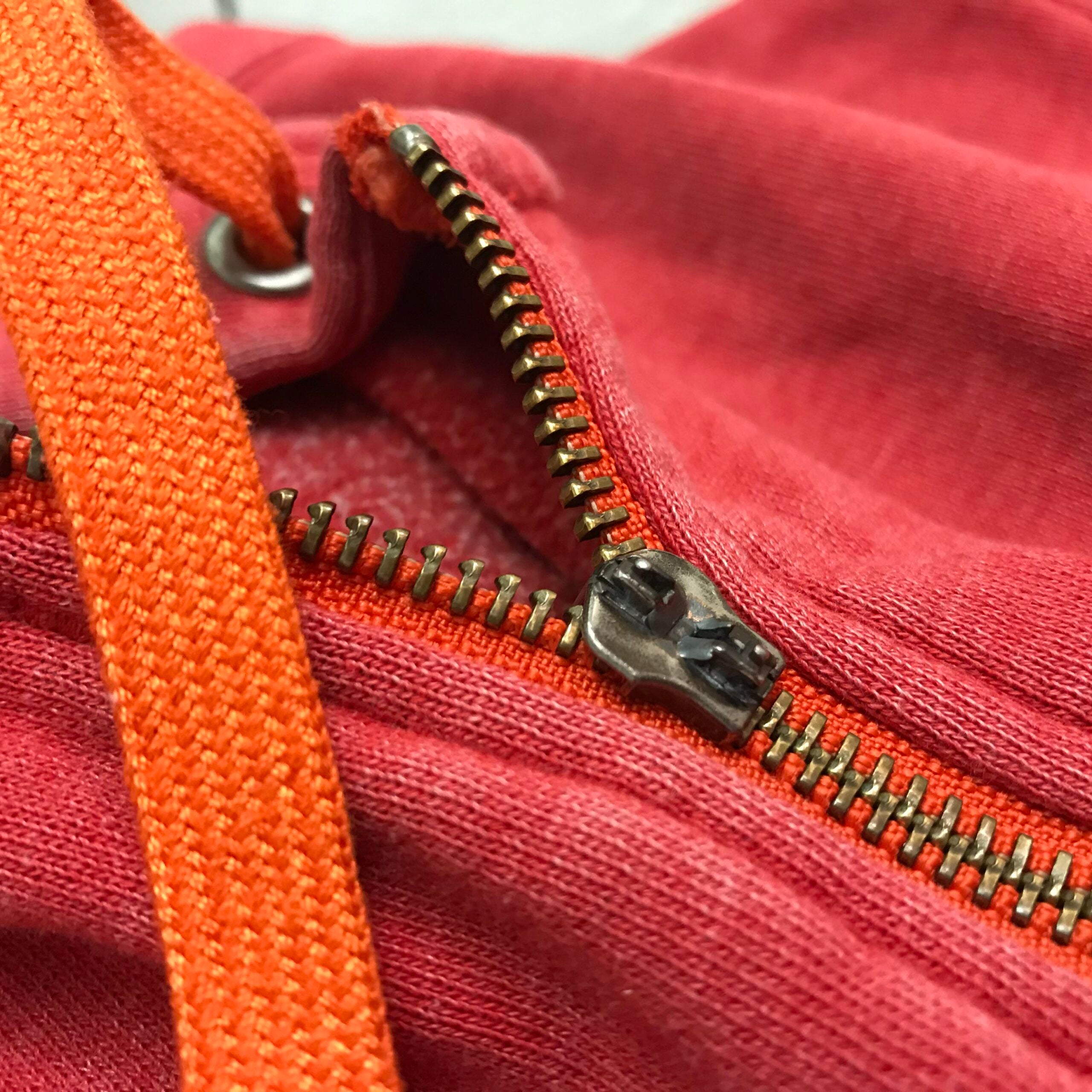 Close-up of a partially open brass zipper on a red fabric, with an orange drawstring crossing over the zipper.