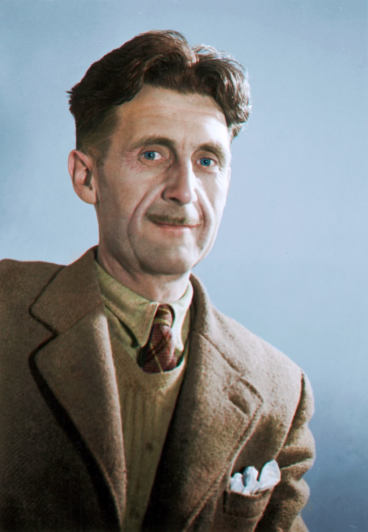 A man with short brown hair and blue eyes, wearing a brown suit jacket, beige sweater, collared shirt, and plaid tie, sits against a plain blue background, looking calmly at the camera.
