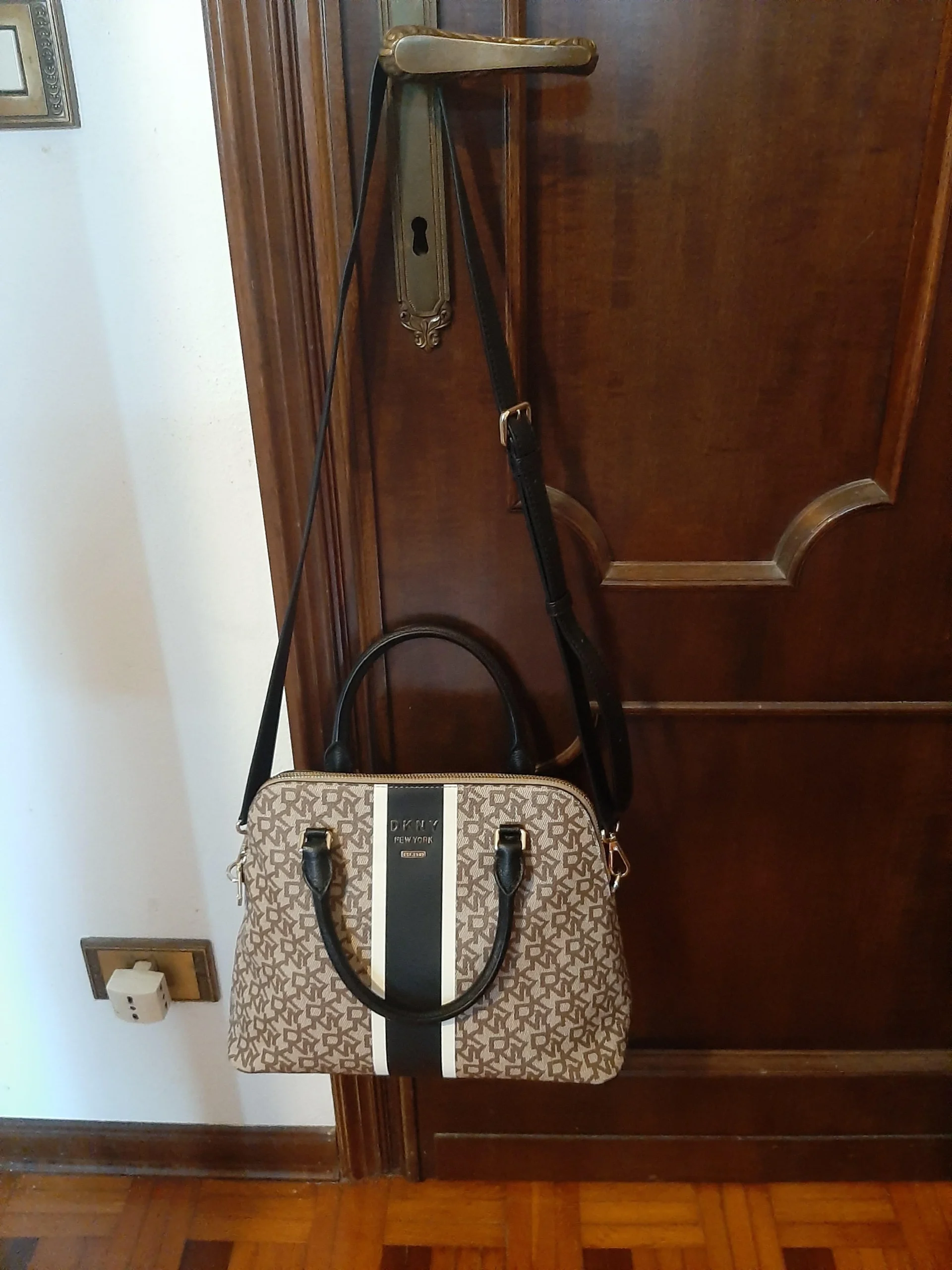 A brown patterned handbag with black handles and a shoulder strap hangs from a brass door handle on a dark wooden door. The bag features a white and black vertical stripe and sits above a wooden floor.