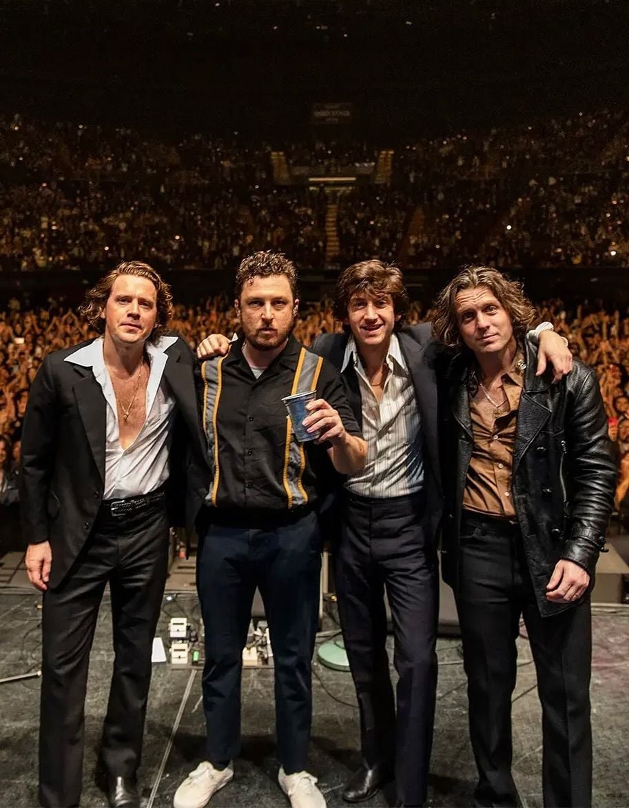Four men stand on stage with their arms around each other, facing the camera, in front of a large cheering crowd in a concert venue. They are dressed in retro-inspired outfits and look relaxed and happy.