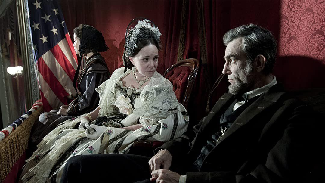 Three people in 19th-century attire sit in a theater box decorated with red drapes and an American flag; a woman in a white dress looks intently at a bearded man in a suit, while another woman sits beside them.