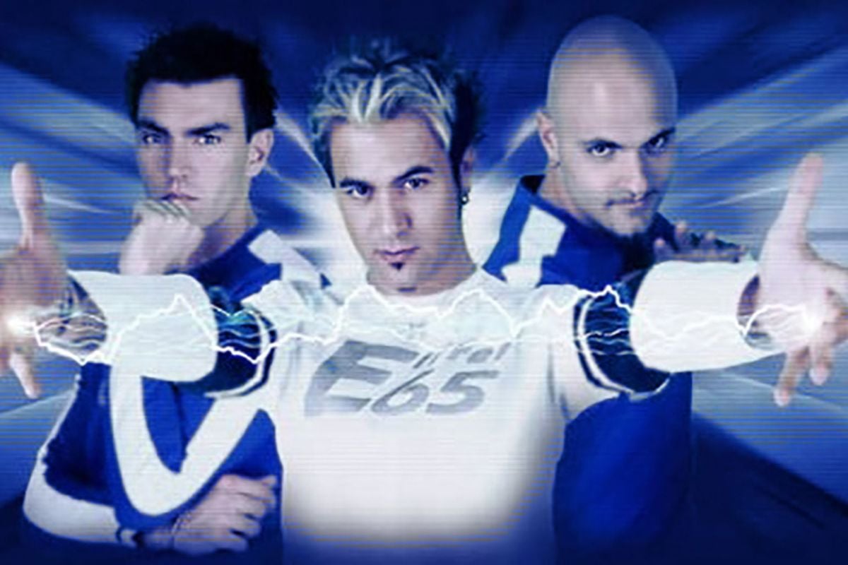Three men pose confidently with intense expressions against a blue, digitally-styled background. The man in front extends his hands forward, with lightning effects, wearing a white shirt that says "Eiffel 65.