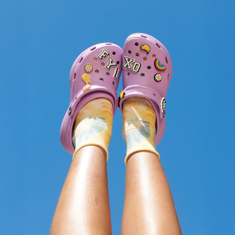 A person points their legs upward against a bright blue sky, wearing pastel tie-dye socks and light purple Crocs decorated with colorful pins and charms.