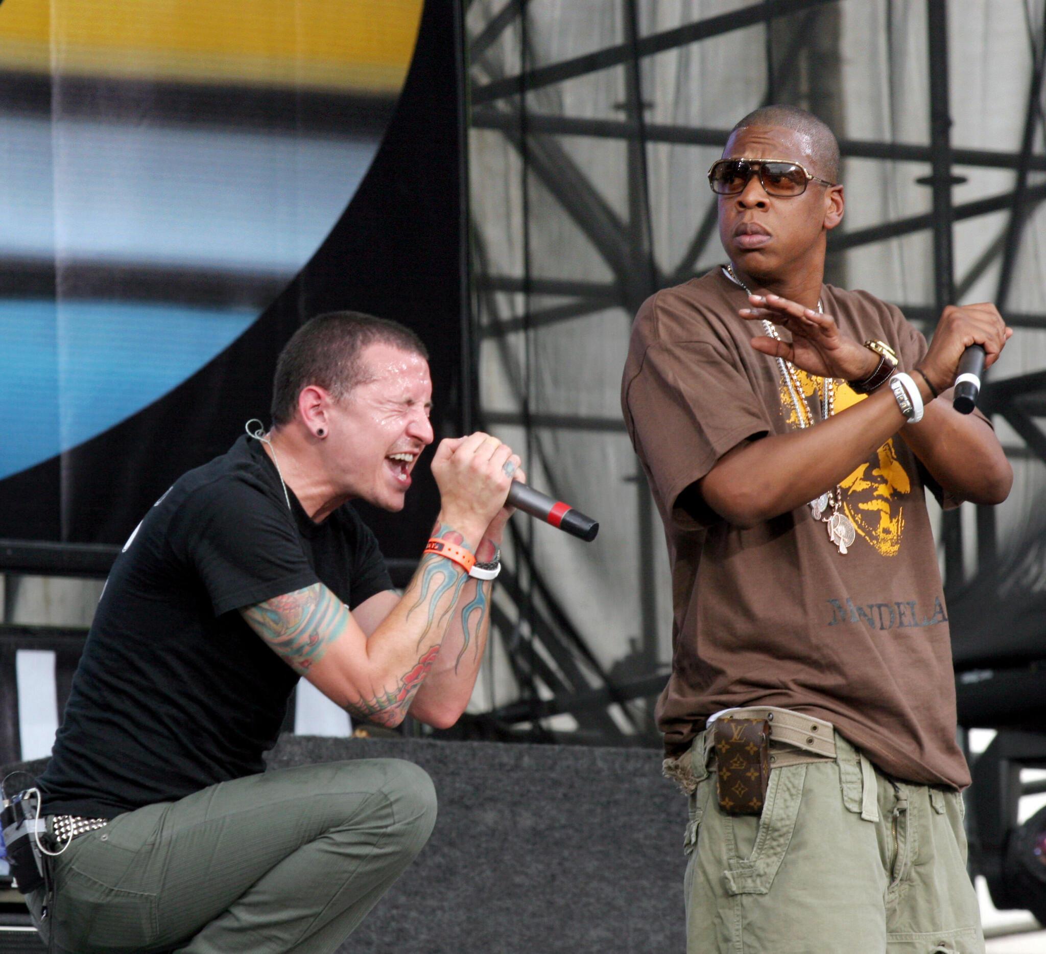 Two male performers are on stage. One is kneeling and singing passionately into a microphone, while the other stands beside him, holding a microphone and looking to the side, wearing sunglasses and a brown T-shirt.