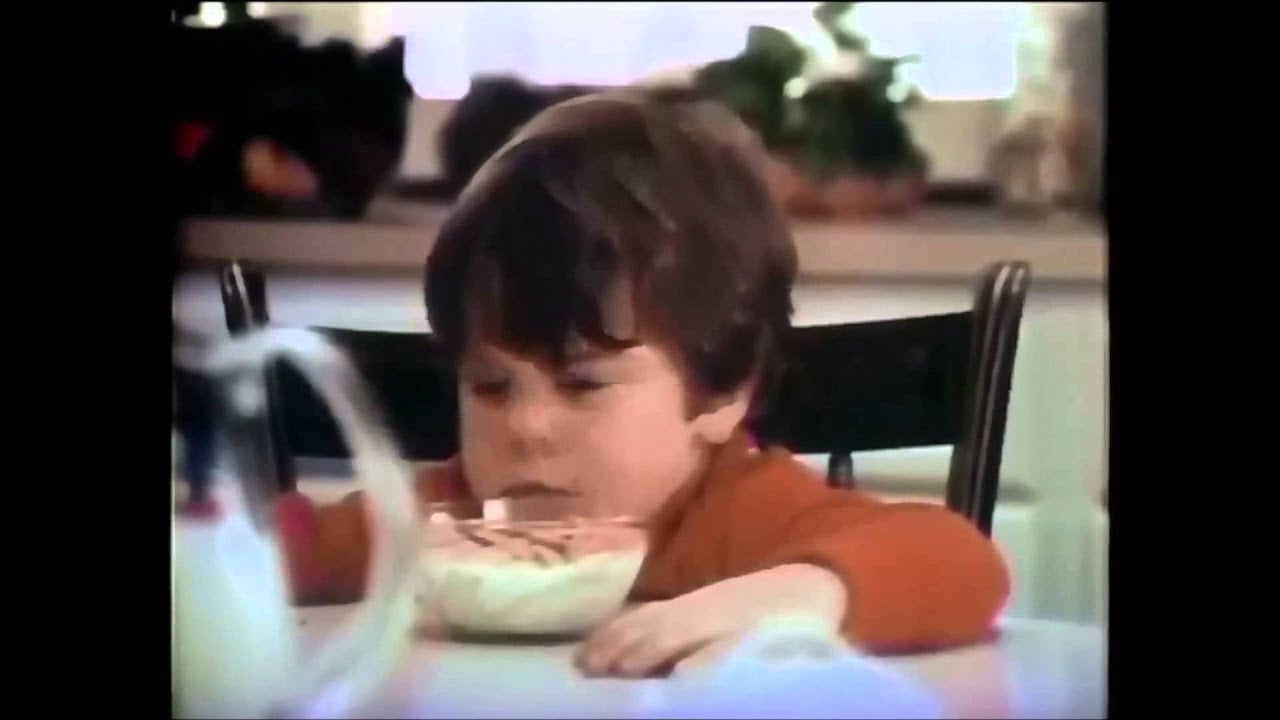 A young boy with brown hair sits at a table, looking down at a bowl of cereal. The background shows a bright kitchen with plants on the windowsill.