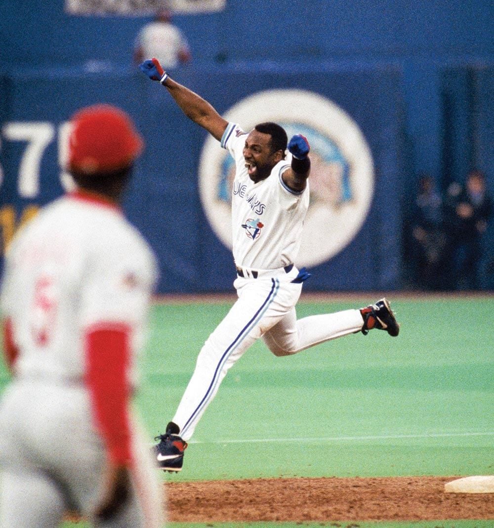 A baseball player in a white Toronto Blue Jays uniform jumps with arms raised in celebration on the field, while a player in a red uniform stands in the foreground.