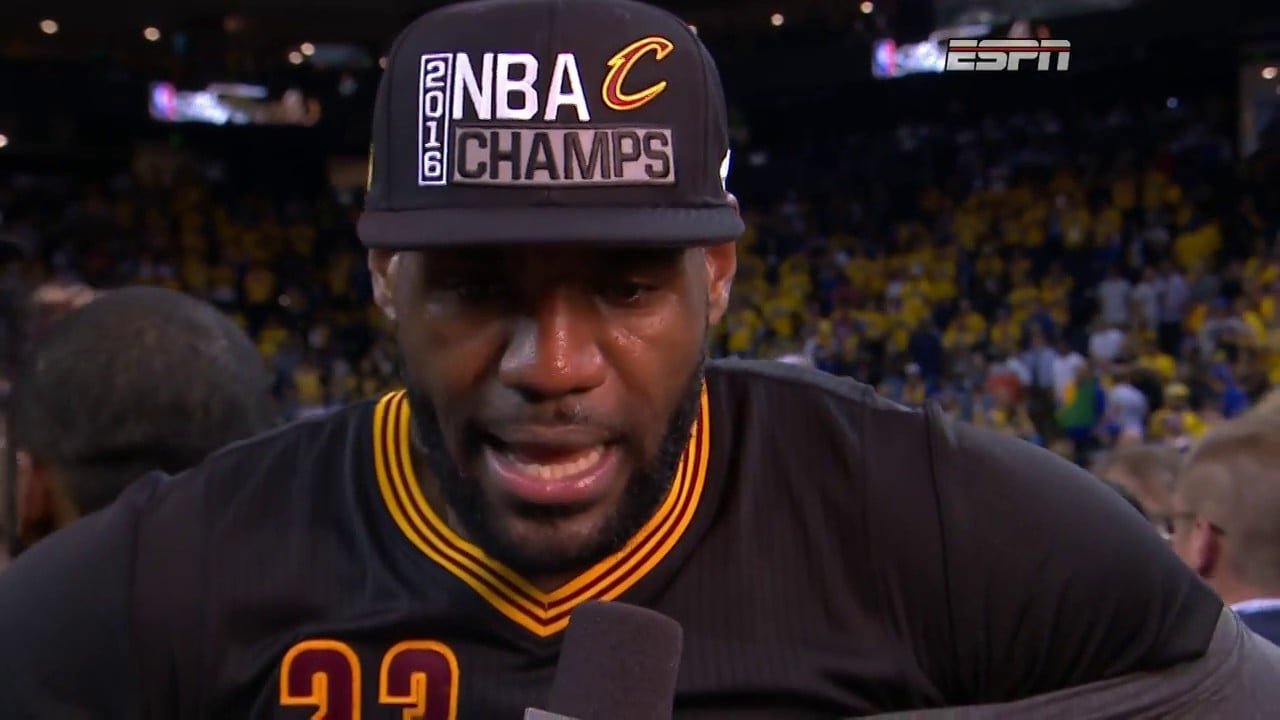 LeBron James in a black Cleveland Cavaliers jersey and a "2016 NBA Champs" hat speaking to the media after winning the 2016 NBA Finals, with a crowd in the background.