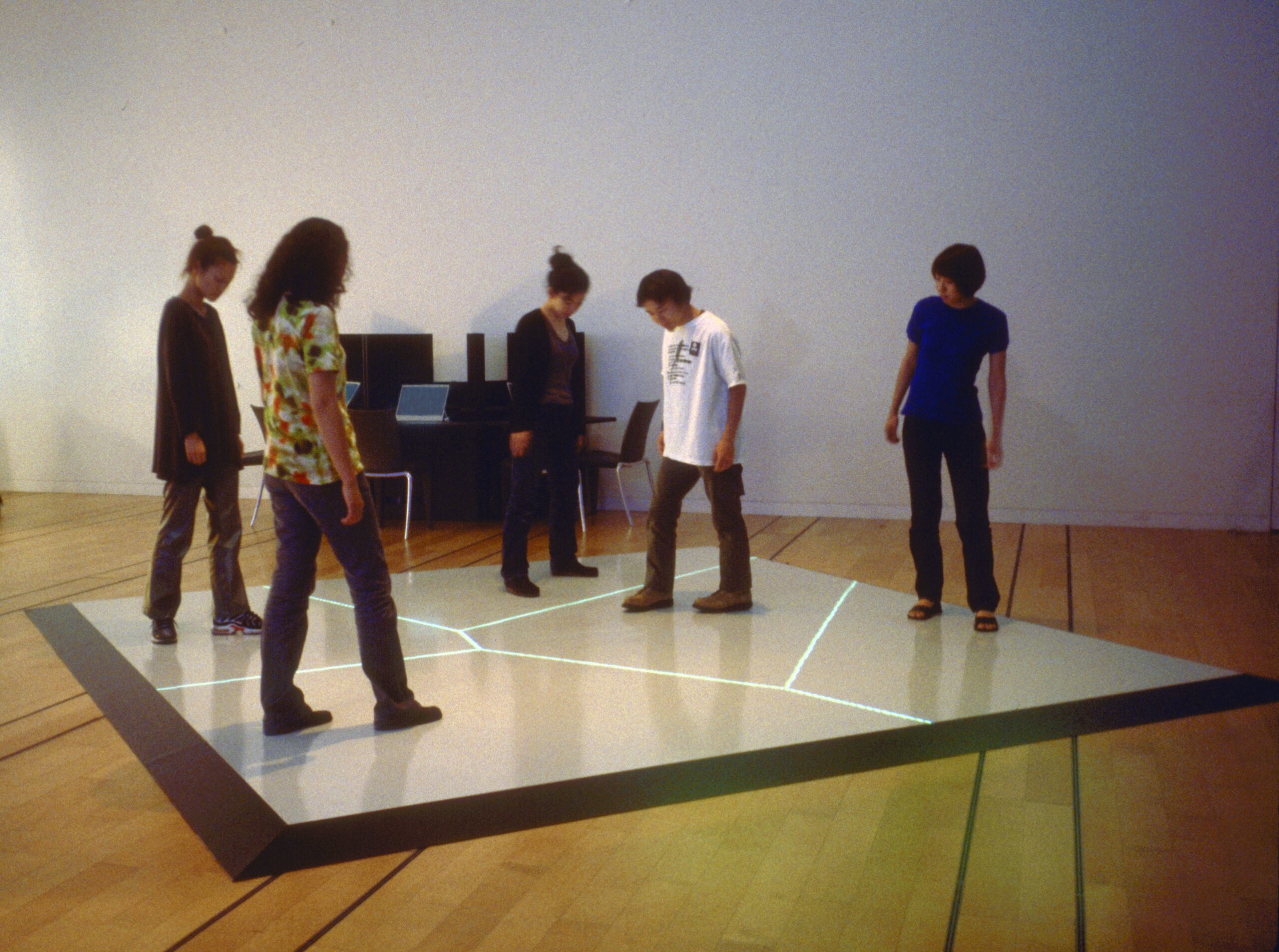 Five people stand on a large, interactive floor display with glowing lines, inside a minimalist room with wood floors, white walls, and chairs and a computer setup in the background.