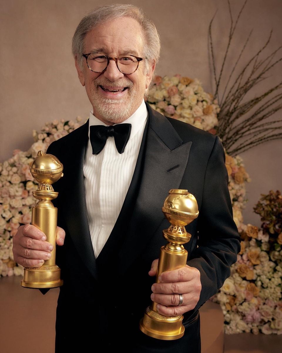An older man in a tuxedo smiles while holding two Golden Globe awards, standing in front of a floral backdrop.