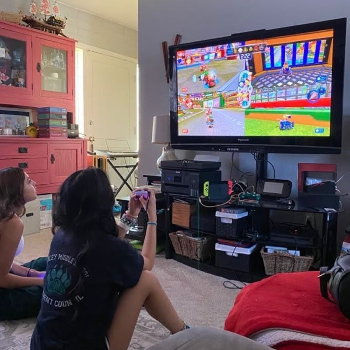 Two people sit on the floor in a living room, playing a colorful video game on a large TV screen. Gaming consoles and accessories are on the TV stand, with shelves and a red cabinet in the background.