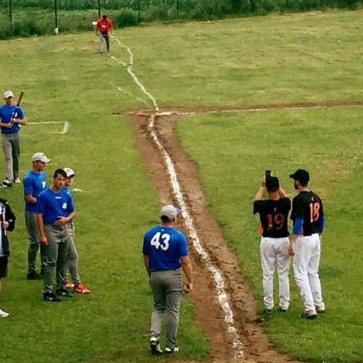 A baseball field with a crooked white foul line. Two players in black jerseys, numbered 19 and 18, take a selfie, while others in blue shirts stand nearby on the grass. A person walks near the outfield fence.