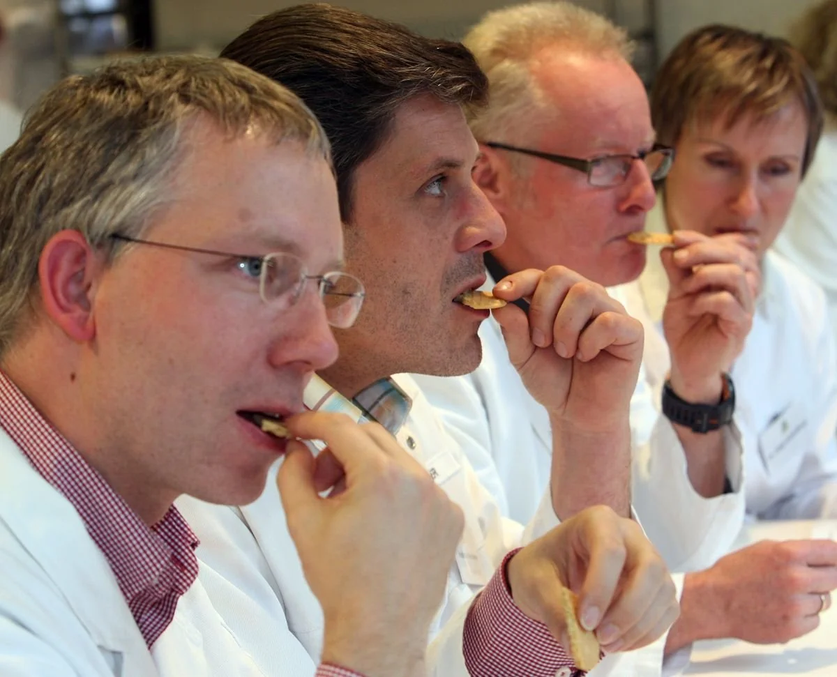 Four people in white coats sit in a row, closely tasting crackers or biscuits, appearing focused as they evaluate the food.