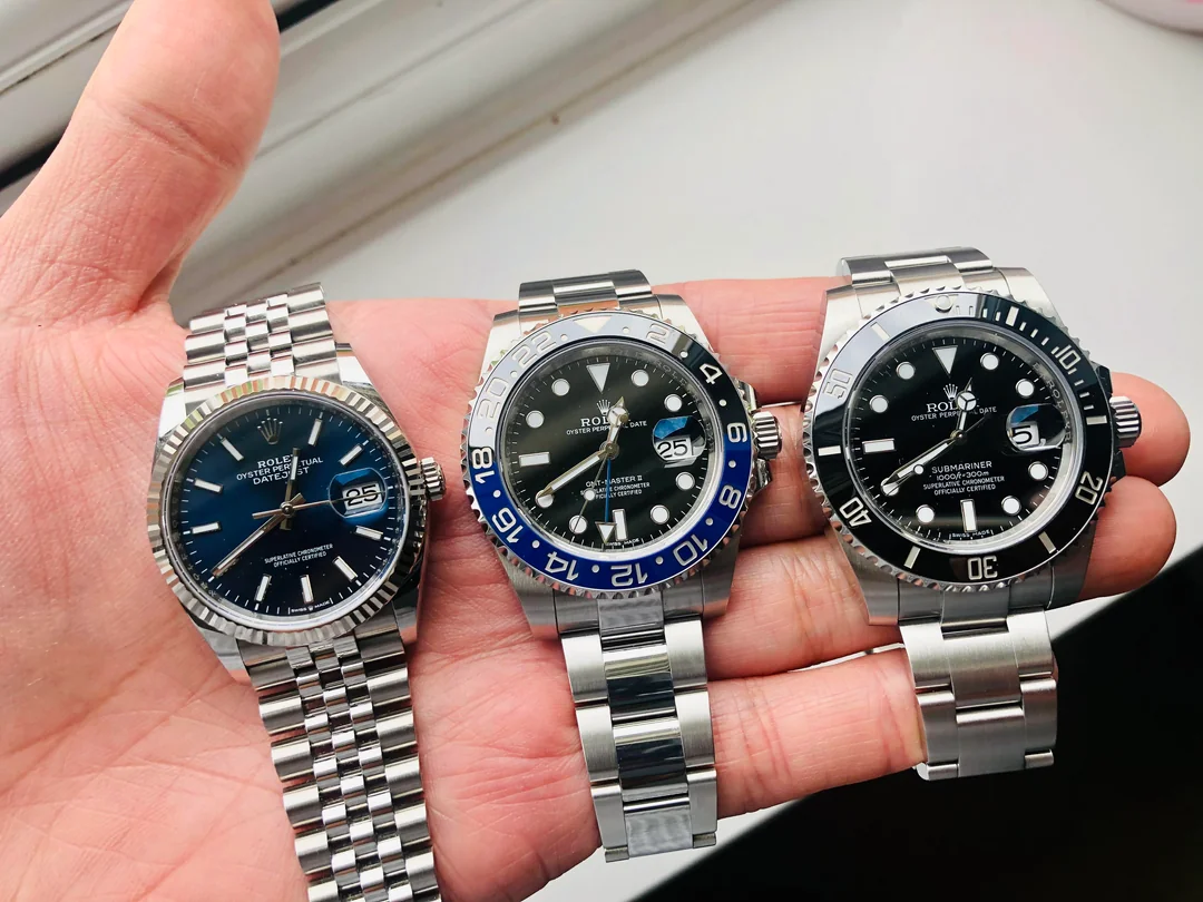 A hand holds three luxury Rolex watches with metal bands, each displaying the date and time. The left watch has a blue face, the middle has a black and blue bezel, and the right has a black face and bezel.