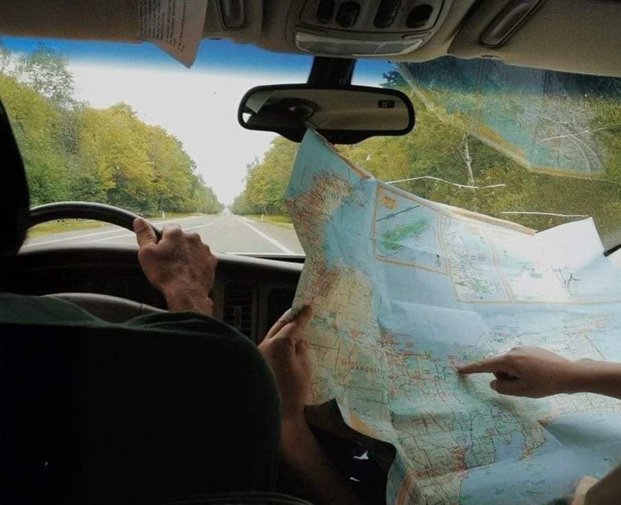 Two people sit in a car; one is driving while the other holds a large unfolded map, pointing at a location. The road ahead is lined with trees and stretches into the distance.