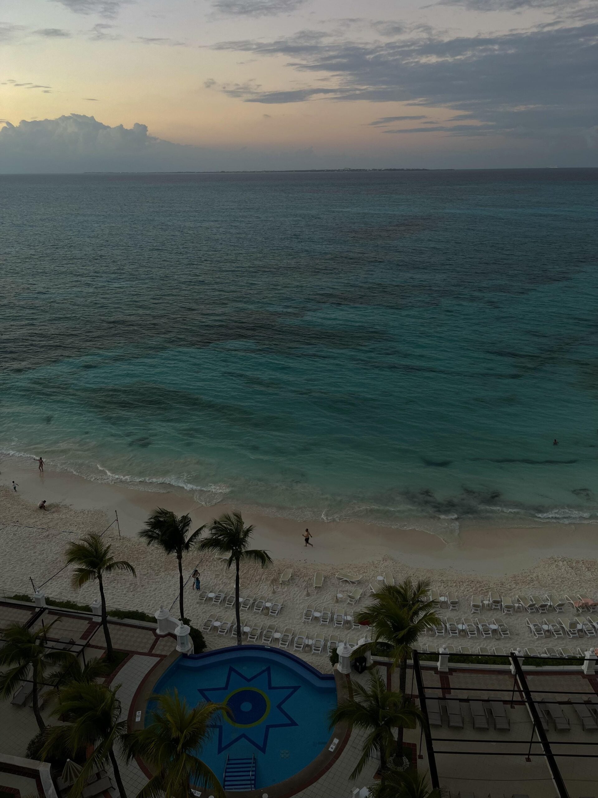 View from above of a beach at sunset with turquoise ocean waves, white sand, several palm trees, and a circular pool with a blue star design. A few people are walking along the shore, and lounge chairs line the beach.