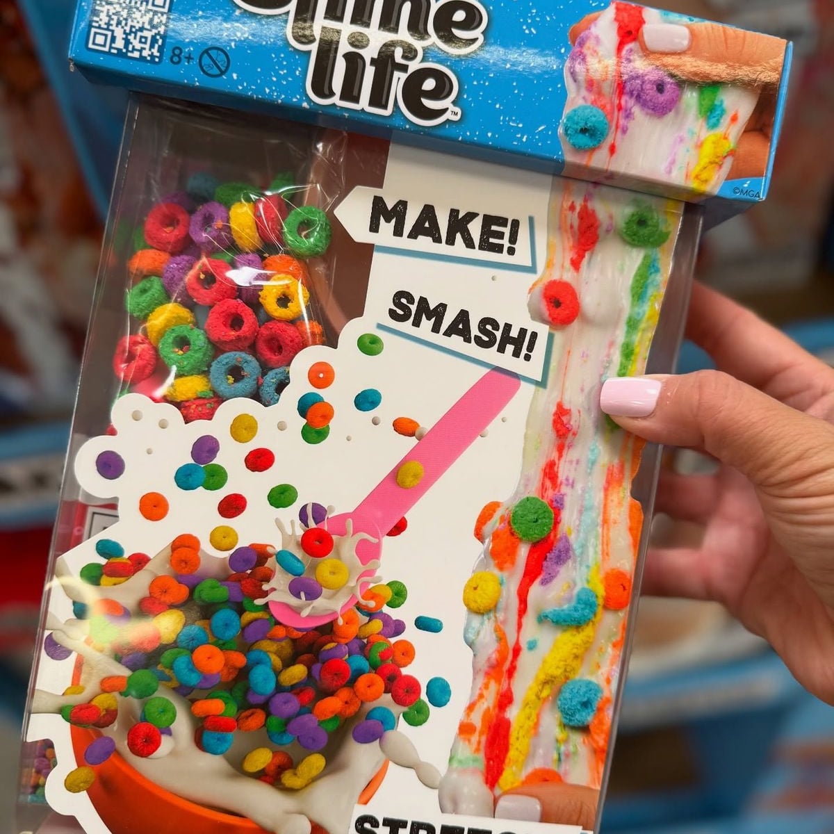 A hand holds a colorful Slime Life kit box featuring images of rainbow cereal pieces, white slime, and a pink spoon, with the words "MAKE!" and "SMASH!" on the packaging.