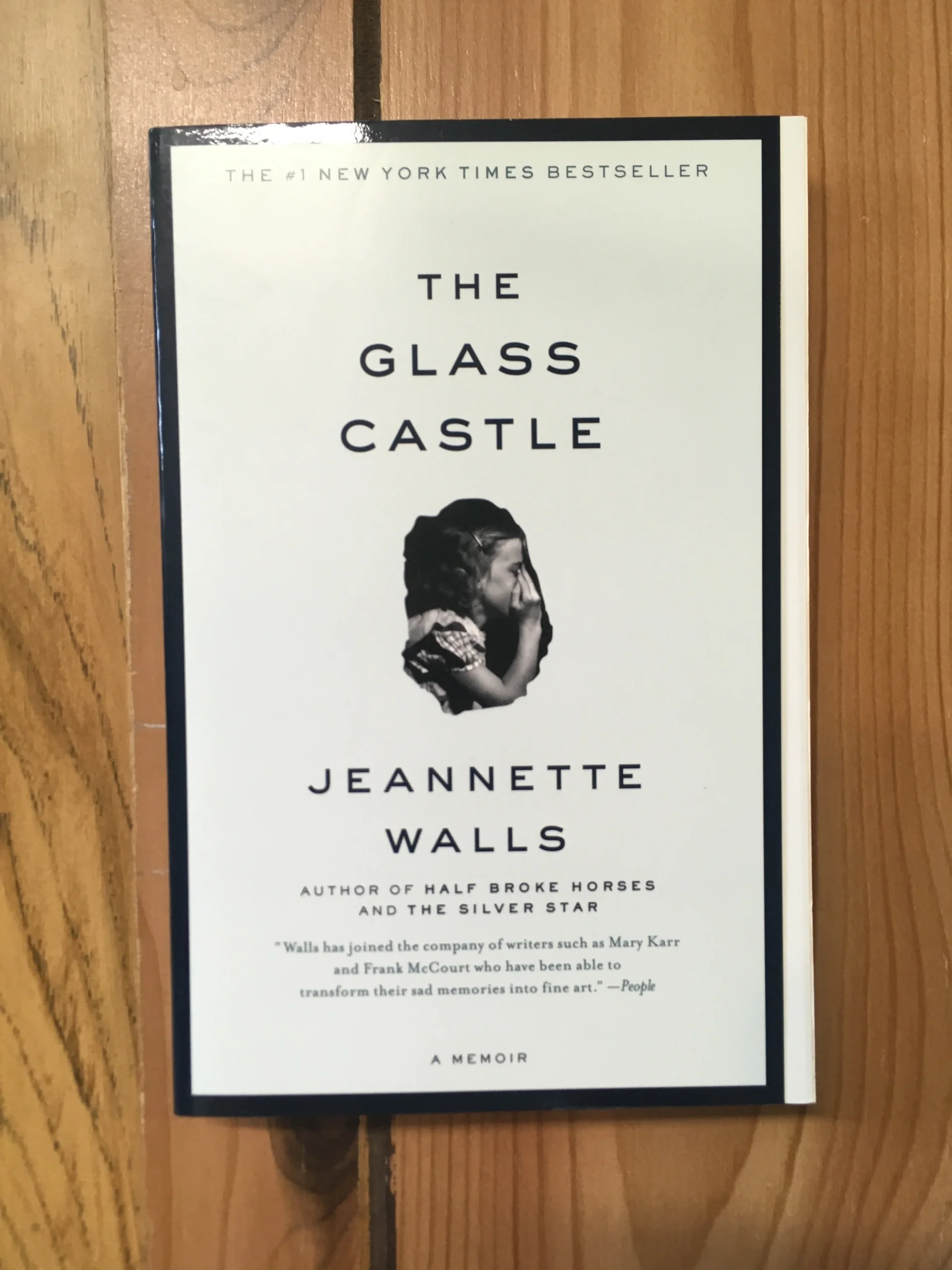 The book "The Glass Castle" by Jeannette Walls is placed on a wooden surface. The cover features a black-and-white profile photo of a young girl and bold black text on a white background.