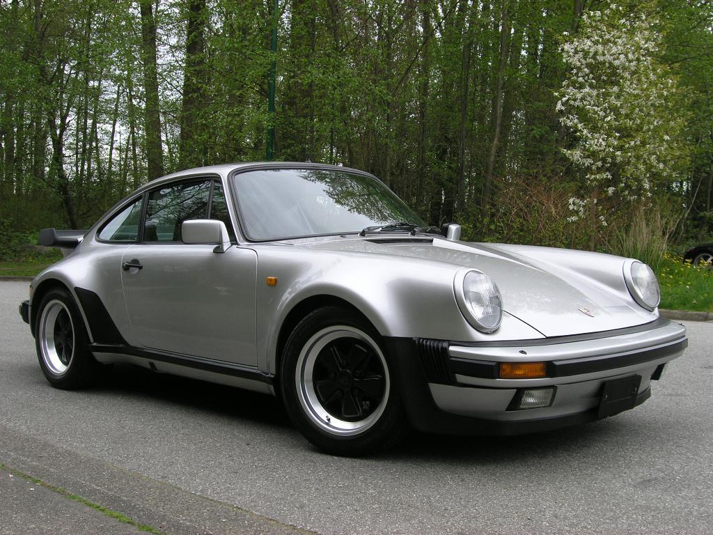 A silver classic Porsche 911 sports car is parked on a road with trees and greenery in the background. The car features a wide body, black trim, and a distinctive rear spoiler.