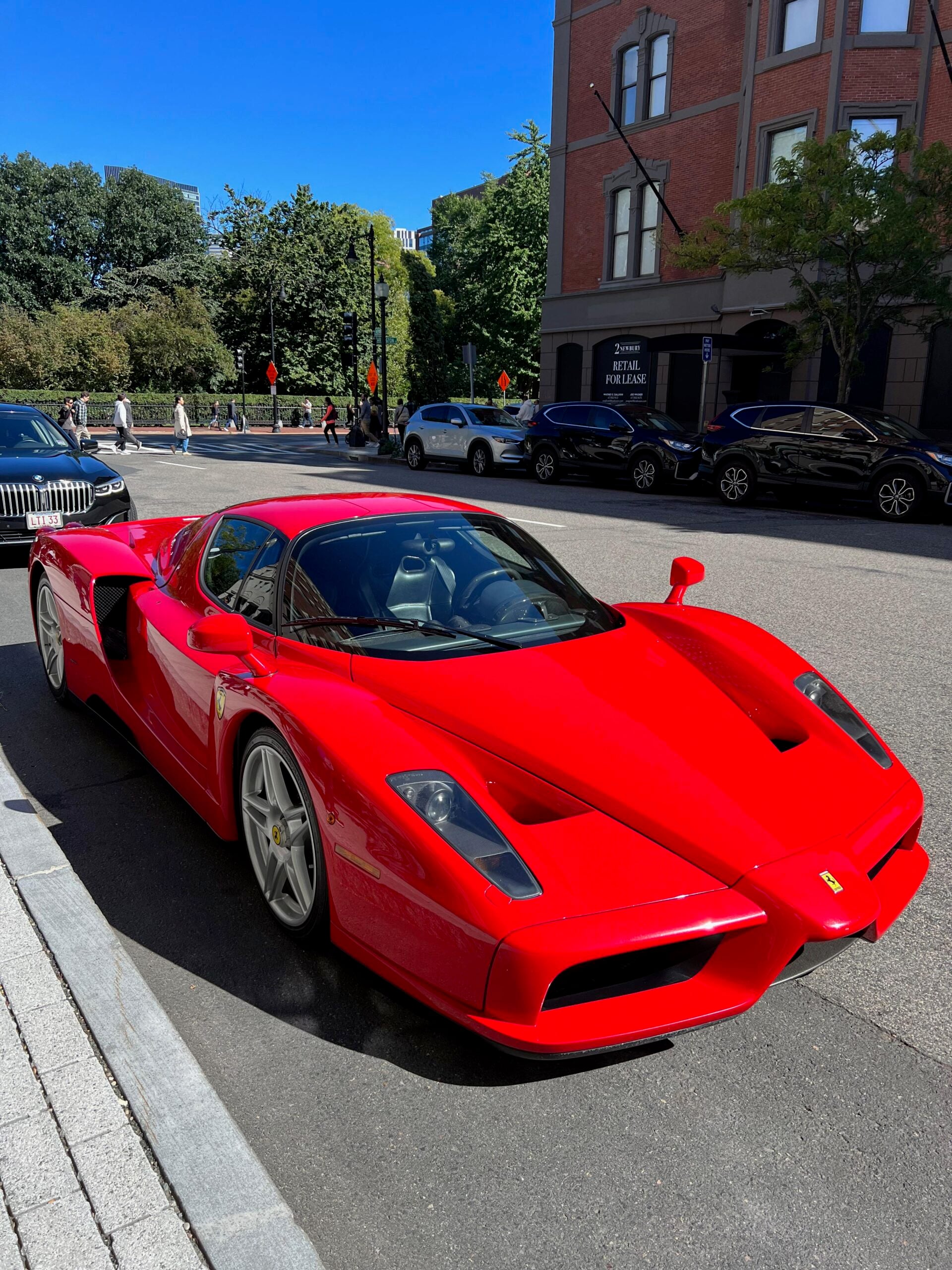 A bright red Ferrari sports car is parked on a city street near a brick building, with trees, parked cars, and pedestrians visible in the background on a sunny day.