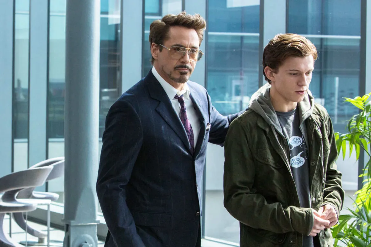 A man in a suit and sunglasses places his hand on the shoulder of a younger man in a jacket and hoodie, as they stand in a modern building with large windows and sunlight.