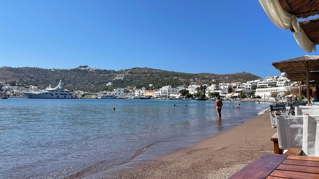 A sandy beach with calm, clear water, a few people swimming and walking, white buildings and hills in the background, and yachts anchored near the shore under a clear blue sky. Outdoor seating is visible on the right.