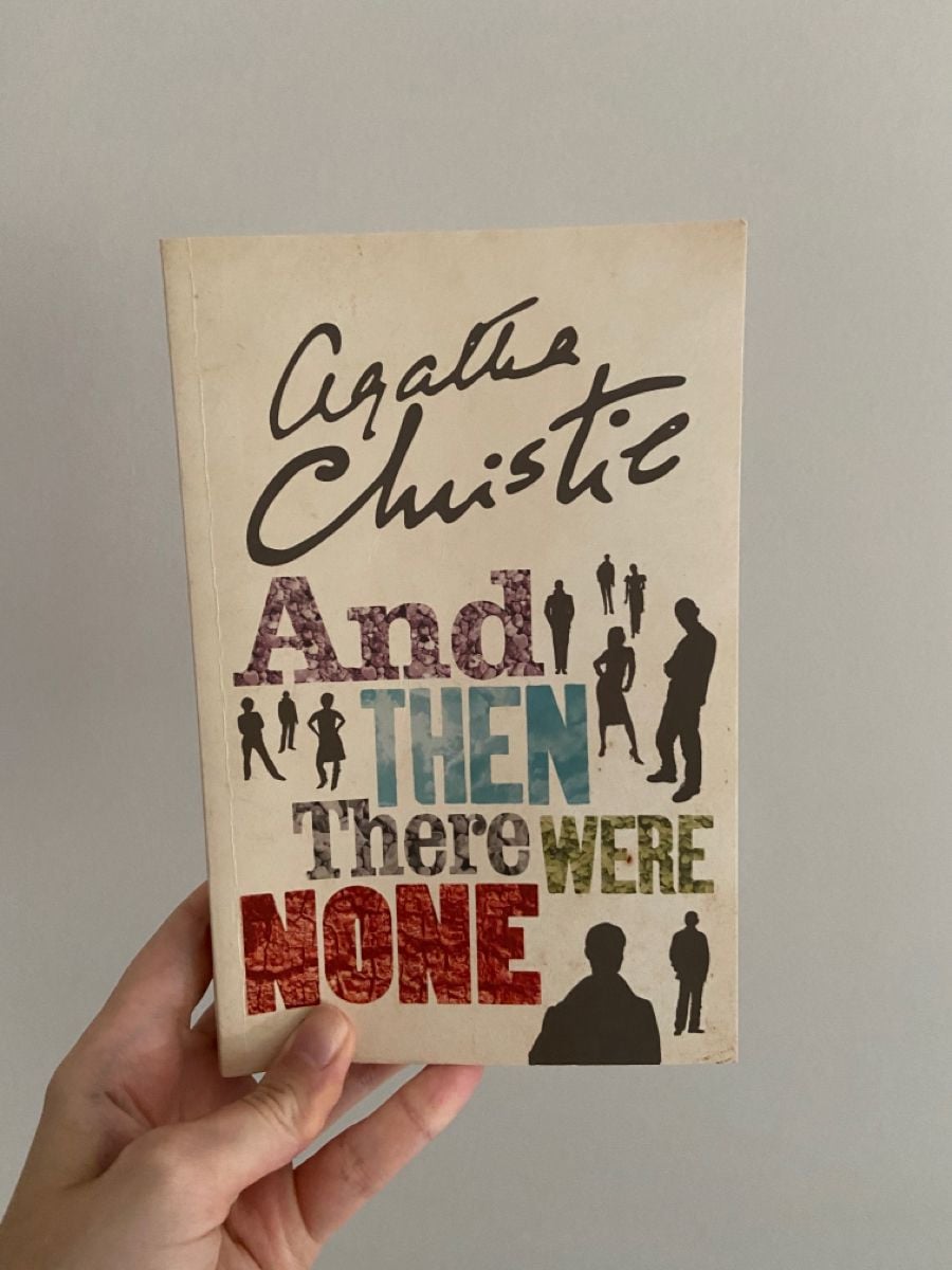 A hand holds a book titled "And Then There Were None" by Agatha Christie. The cover features silhouettes of people in various colors and sizes against a plain background.