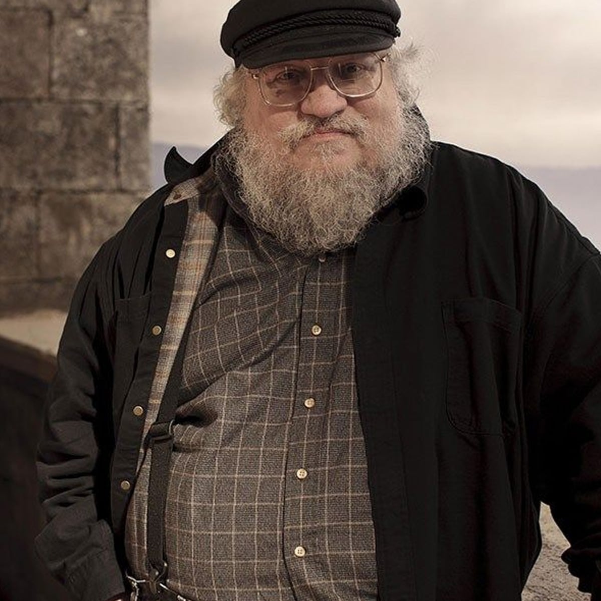 An older man with a white beard and glasses stands outdoors, wearing a black cap, dark shirt, and plaid button-up. He is looking at the camera with a neutral expression. Stonework and an overcast sky are in the background.