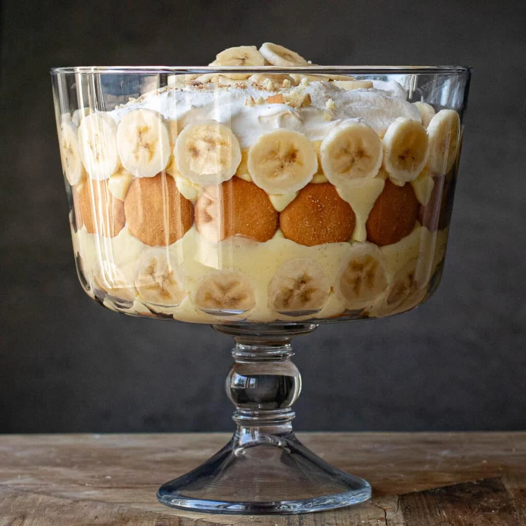 A glass trifle bowl filled with layers of sliced bananas, vanilla wafers, creamy pudding, and whipped topping, arranged in an elegant and appetizing presentation on a wooden surface.