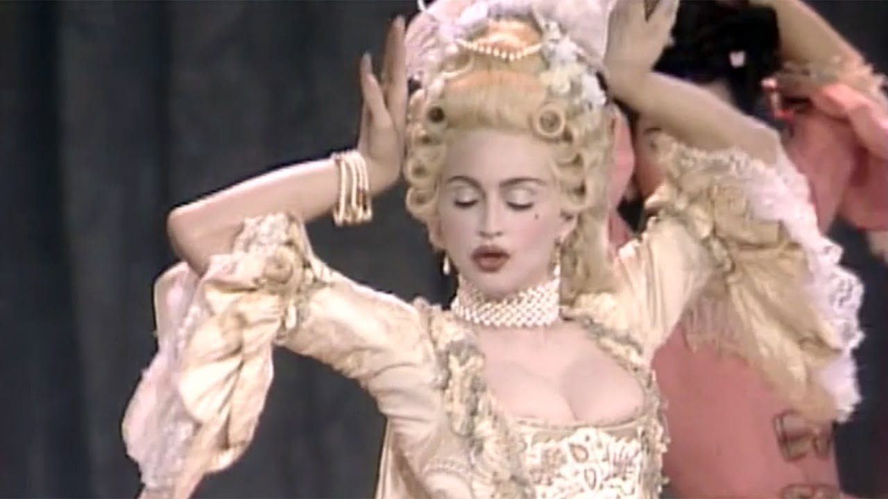 A woman dressed in ornate 18th-century-style clothing and a tall powdered wig poses with her hands raised. She wears pearls, lace, and makeup, evoking a Marie Antoinette-inspired look.