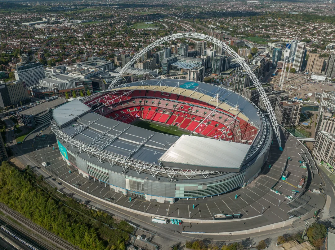 Aerial view of Wembley Stadium in London, featuring its iconic arch, red seating, and surrounding city buildings on a clear day.