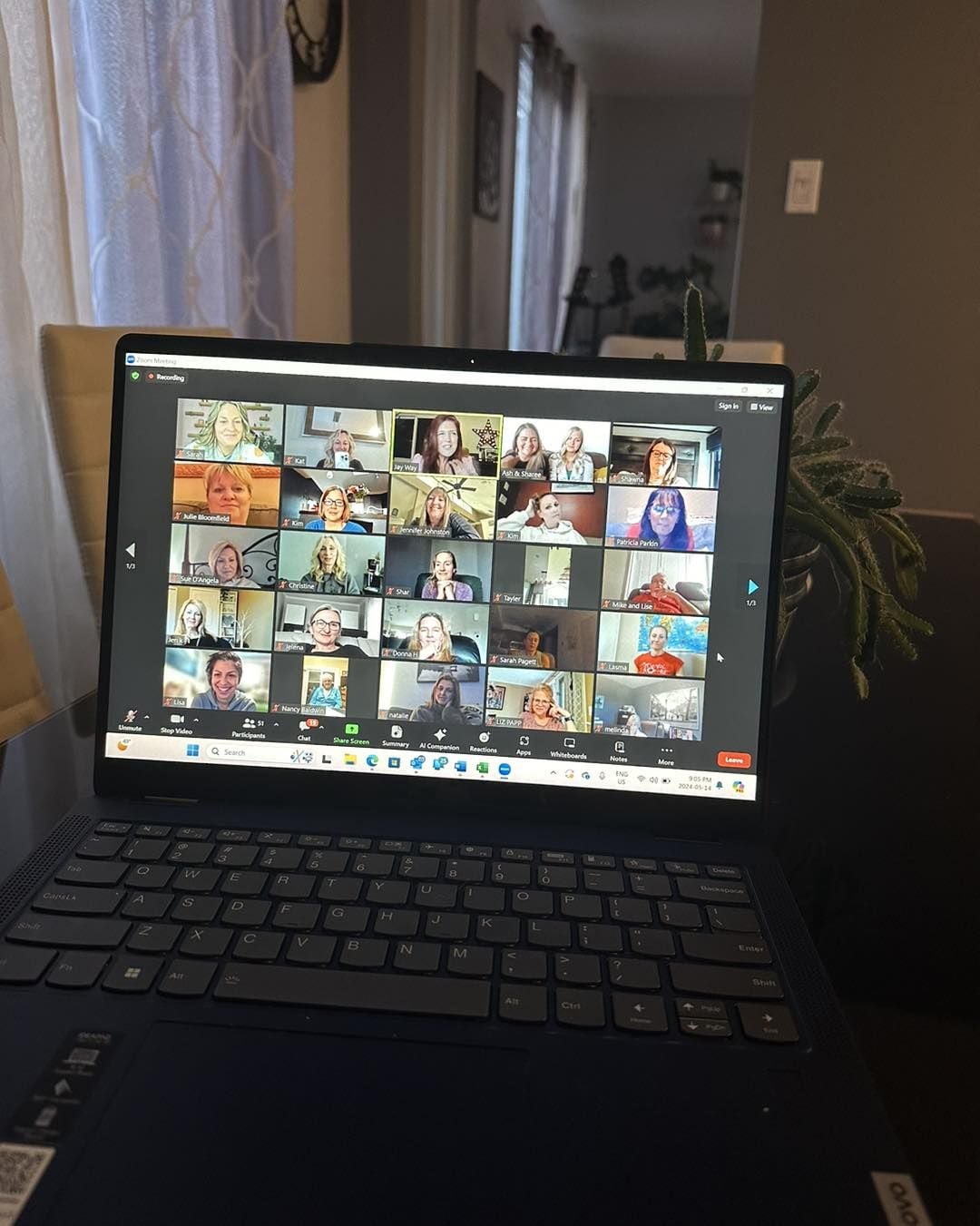 A laptop displays a virtual meeting with over 20 participants on a video call grid. The laptop sits on a table in a well-lit room with sheer curtains and houseplants in the background.