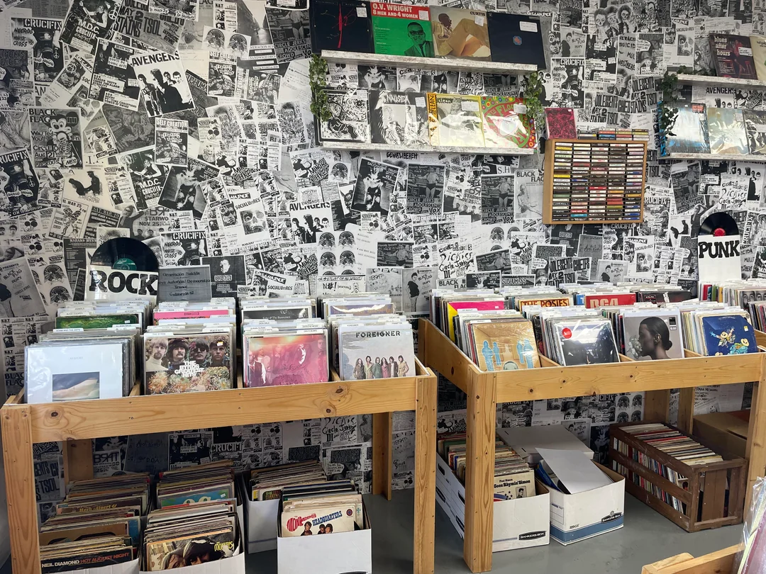 An indie record store interior with wooden bins full of vinyl records, boxes of records on the floor, and shelves of tapes. The wall is covered in black-and-white band posters and album covers, creating a vintage, punk-rock vibe.