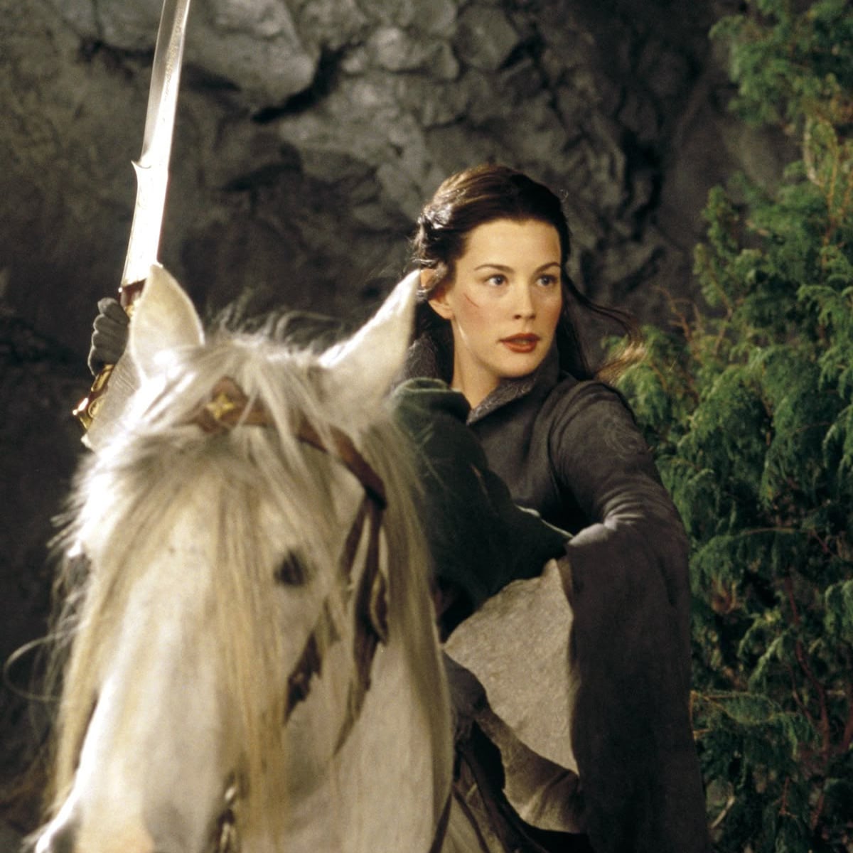 A woman with dark hair wearing medieval-style clothing rides a white horse, holding a raised sword. She looks alert and determined, with greenery and a rocky background behind her.